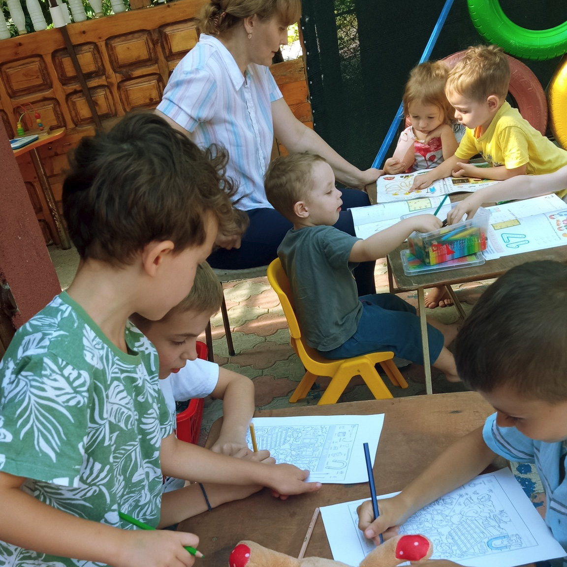 Preșcolari colorează fișe sub îndrumarea educatoarei, în aer liber