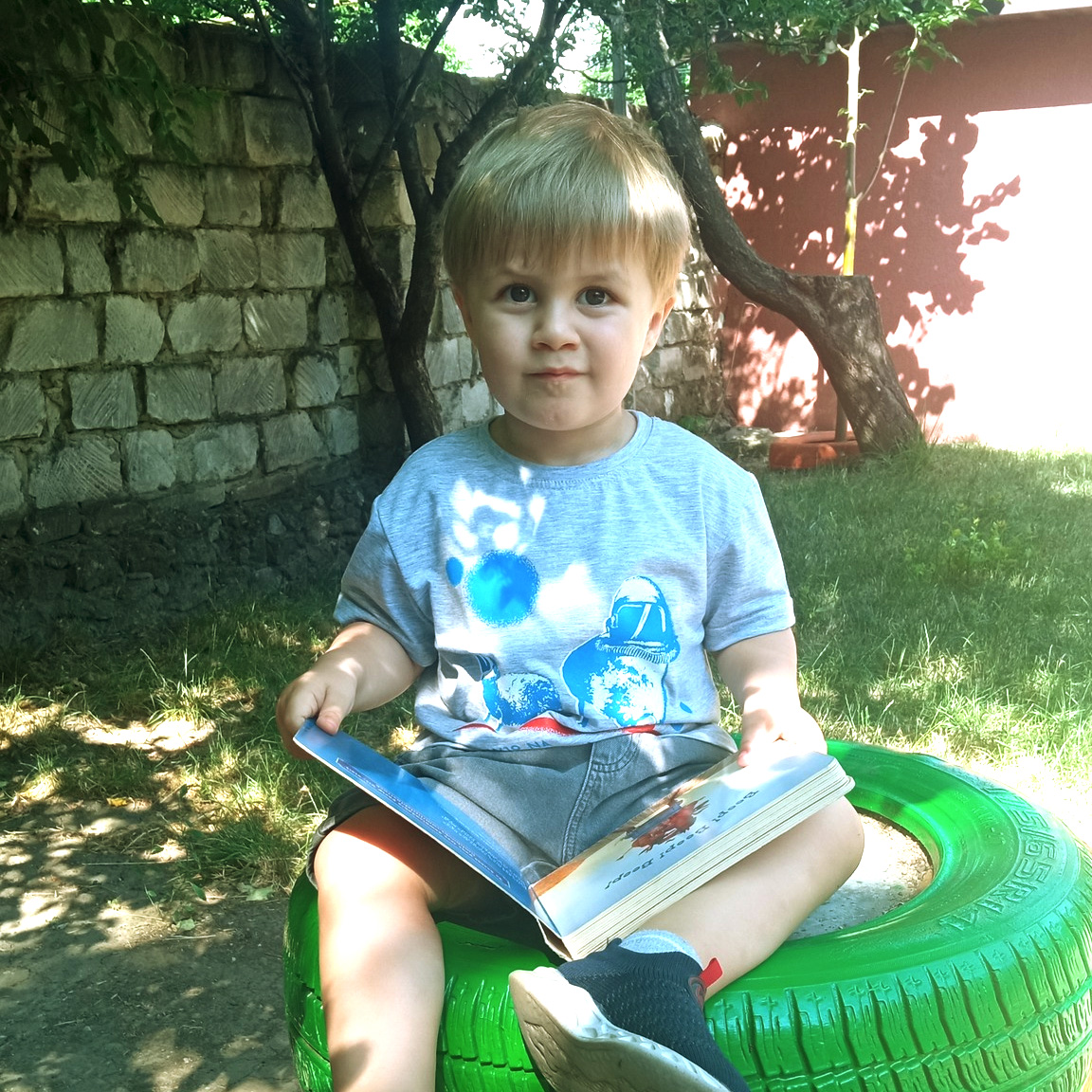 Copil citește o carte ilustrată, în curtea grădiniței