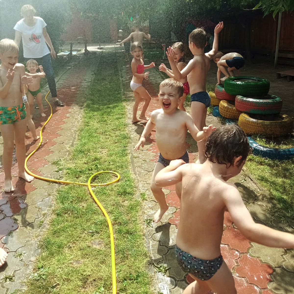 Joacă de vară: copii zburdă fericiţi prin stropii de apă din furtun